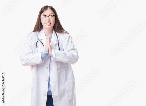 Young Chinese doctor woman over isolated background begging and praying with hands together with hope expression on face very emotional and worried. Asking for forgiveness. Religion concept.