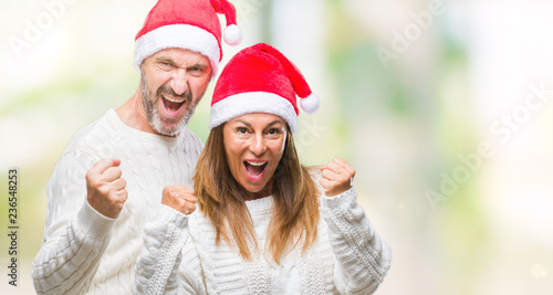 Middle age hispanic couple wearing christmas hat over isolated background very happy and excited doing winner gesture with arms raised, smiling and screaming for success. Celebration concept.
