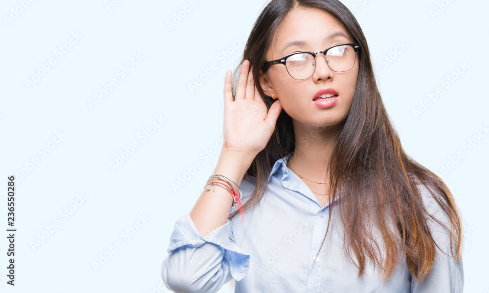Young asian business woman wearing glasses over isolated background smiling with hand over ear listening an hearing to rumor or gossip. Deafness concept. Young asian business woman wearing glasses over isolated background smiling with hand over ear listening an hearing to rumor or gossip. Deafness concept.