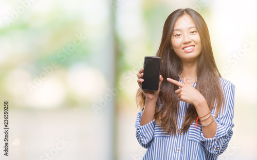 Young asian woman showing blank screen of smartphone over isolated background very happy pointing with hand and finger