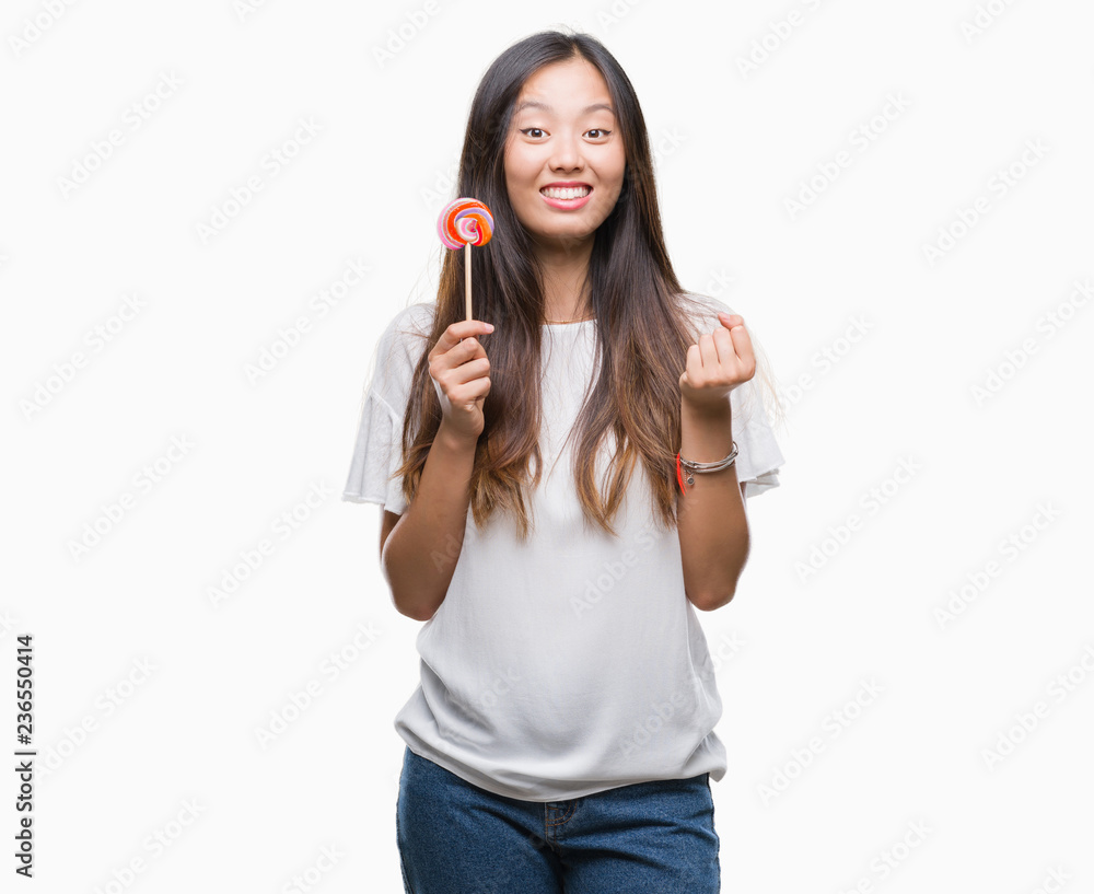 Young asian woman eating lollipop candy over isolated background screaming proud and celebrating victory and success very excited, cheering emotion Young asian woman eating lollipop candy over isolated background screaming proud and celebrating victory and success very excited, cheering emotion