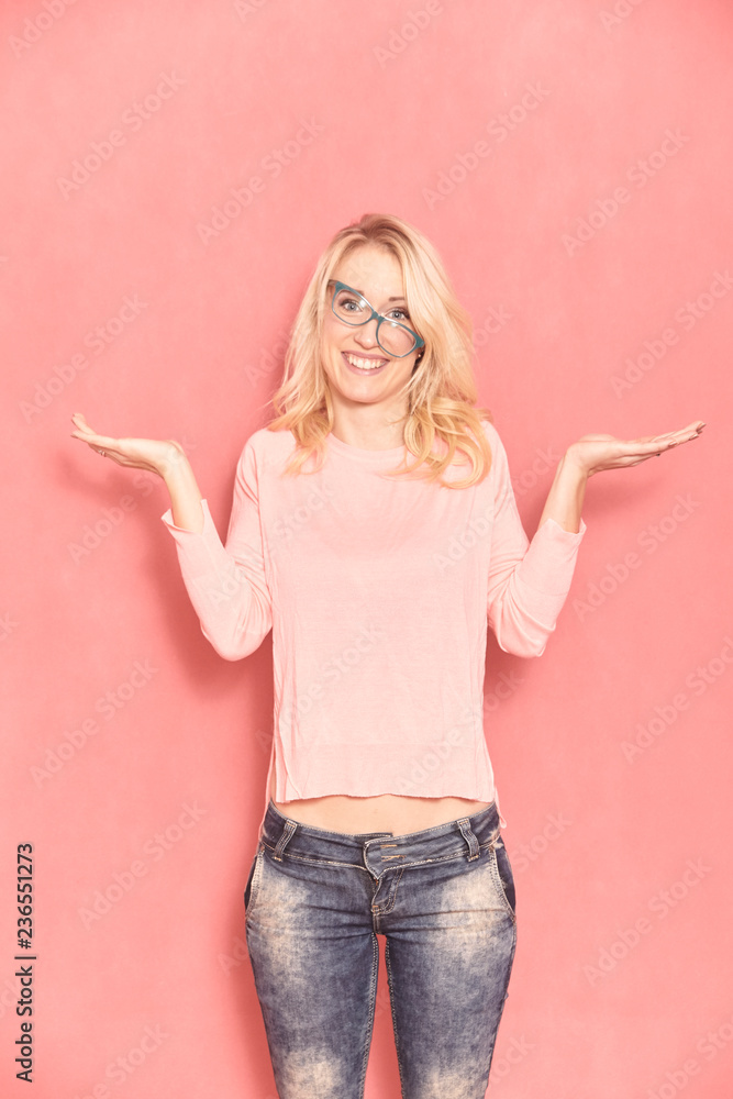one young smiling woman, questioning with her arms raised, posing and looking forward to camera, with her eyeglasses tilted, 20-29 years old, long blond hair. Shot in studio on pink background. - obrazy, fototapety, plakaty 