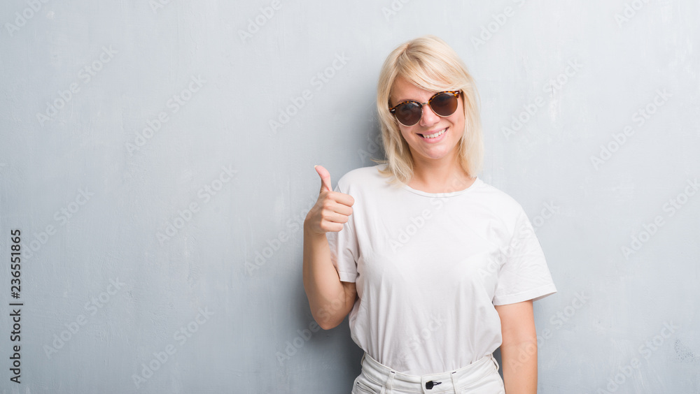 Adult caucasian woman over grunge grey wall wearing sunglasses doing happy thumbs up gesture with hand. Approving expression looking at the camera with showing success. Adult caucasian woman over grunge grey wall wearing sunglasses doing happy thumbs up gesture with hand. Approving expression looking at the camera with showing success.