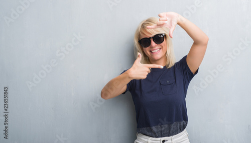 Adult caucasian woman over grunge grey wall wearing sunglasses smiling making frame with hands and fingers with happy face. Creativity and photography concept.