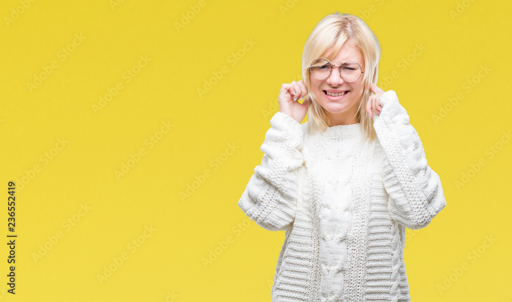 Young beautiful blonde woman wearing winter sweater and glasses over isolated background covering ears with fingers with annoyed expression for the noise of loud music. Deaf concept.
