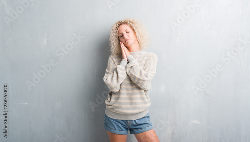 Young blonde woman with curly hair over grunge grey background sleeping tired dreaming and posing with hands together while smiling with closed eyes.