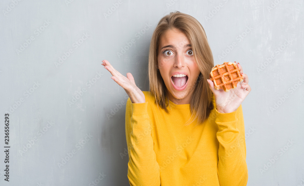 Beautiful young woman over grunge grey wall eating waffle very happy and excited, winner expression celebrating victory screaming with big smile and raised hands