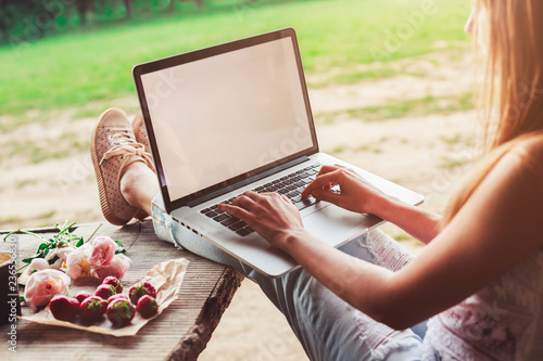 Young woman using and typing laptop computer at rough wooden table with coffee cup, strawberries, bouquet of peonies flowers, smartphone. Freelancer working in outdoor park. Toning photo