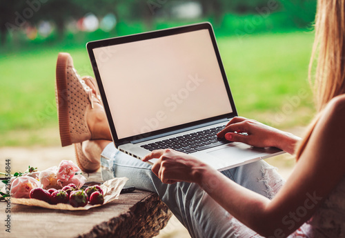 Young woman using and typing laptop computer at rough wooden table with coffee cup, strawberries, bouquet of peonies flowers, smartphone. Freelancer working in outdoor park. Toning photo