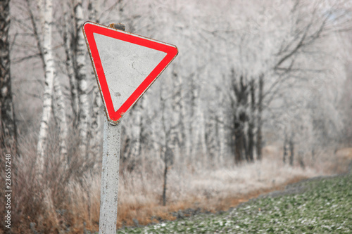 Old, rickety sign give way against the snow-covered forest and field. Road signs, rules of the road. White triangle with a red rim
