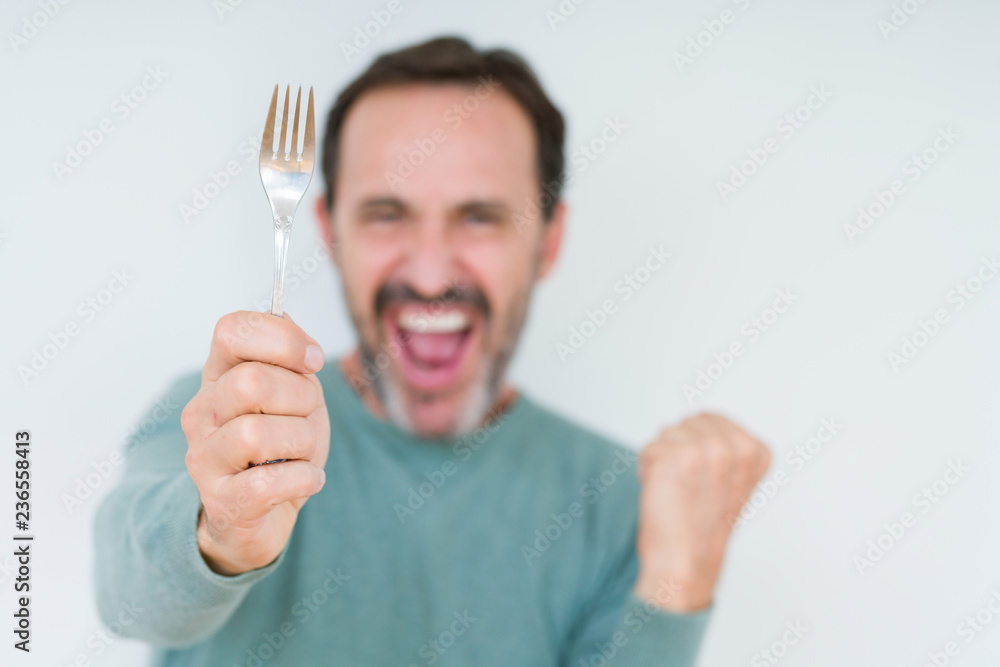 Senior man holding silver fork over isolated background screaming proud and celebrating victory and success very excited, cheering emotion Senior man holding silver fork over isolated background screaming proud and celebrating victory and success very excited, cheering emotion