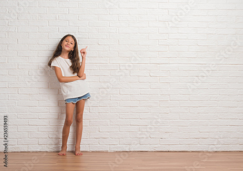 Young hispanic kid stading over white brick wall with a big smile on face, pointing with hand and finger to the side looking at the camera.