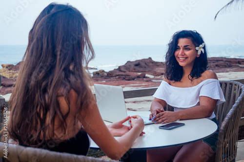 young Indian Asian woman in a business meeting (interview) with a caucasian white woman in tropical paradise beach. multinational handshake and high five successful deal ocean restaurant background © yurakrasil