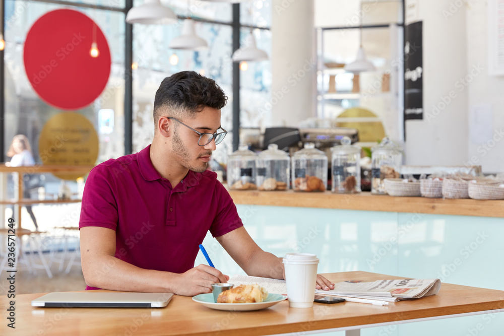 Photo of stylish man with trendy haircut, writes records in notepad, focused in newspaper, drinks takeaway coffee, uses modern laptop computer for freelance work. Hipster guy makes recordings