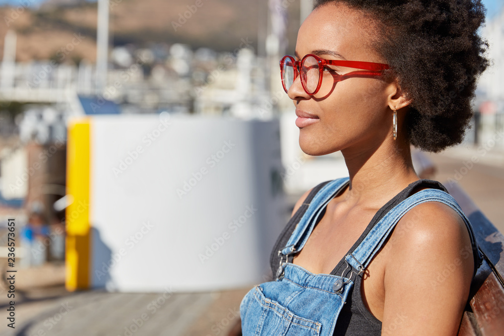 Φωτογραφία Shot of stylish teenage girl with Afro haircut, wears optical glasses for good vision, dressed in denim overalls, models against blurred city background, thinks about new traveling
