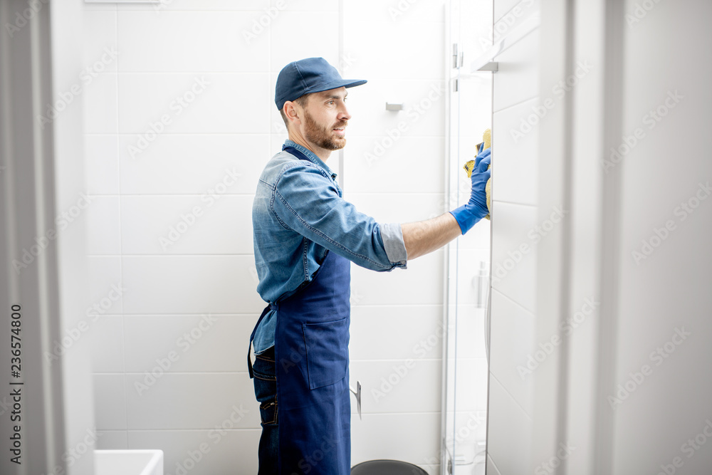Man as professional cleaner wiping the shower door with cotton wiper in the white bathroom Man as professional cleaner wiping the shower door with cotton wiper in the white bathroom