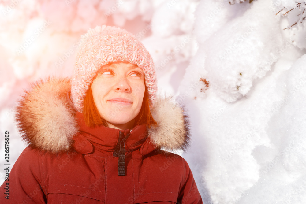 A girl in a pink knitted hat with a pompon and white mittens with a pattern in a warm jacket of copper color with a fur hood stands against the mountain and snow-covered trees A girl in a pink knitted hat with a pompon and white mittens with a pattern in a warm jacket of copper color with a fur hood stands against the mountain and snow-covered trees