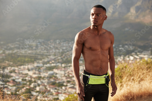 Half length shot of dark skinned Afro man with muscular stomach, goes in for sport regularly, looks aside, wears sportswear, enjoys freedom and healthy lifestyle, has warming up, poses outdoor photo