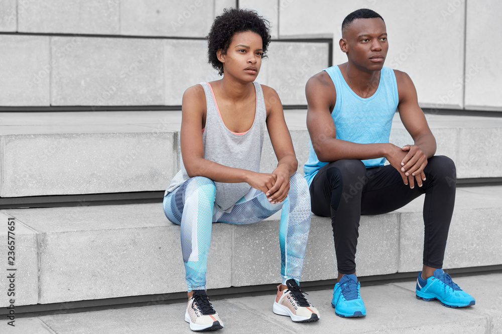 Dark skinned couple dressed in sportswear, comfortable trainers, take break after cardio training, sit at city stairs, focused into distance, being little tired. Girlfriend and boyfriend fond of sport