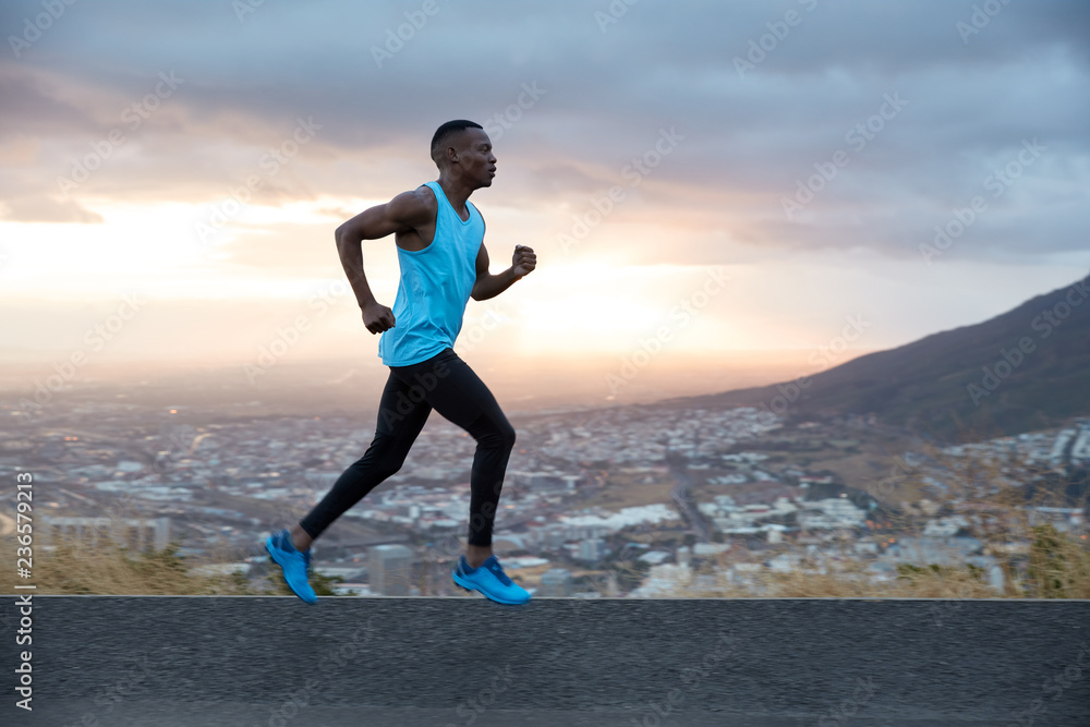 Full length horizontal shot of fast male runner enjoys speed, photographed in motion, runs along road with mountain and city view in background. Running at dawn concept. Strong sportsman outside Full length horizontal shot of fast male runner enjoys speed, photographed in motion, runs along road with mountain and city view in background. Running at dawn concept. Strong sportsman outside