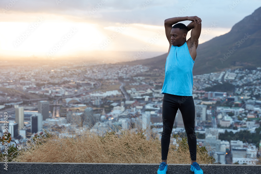 Photo of strong man with good flexibility raises hands above head, does sport exercises in open air at hill against panoramic view with sunrise, town buidling and rocks. Athletic black guy has workout