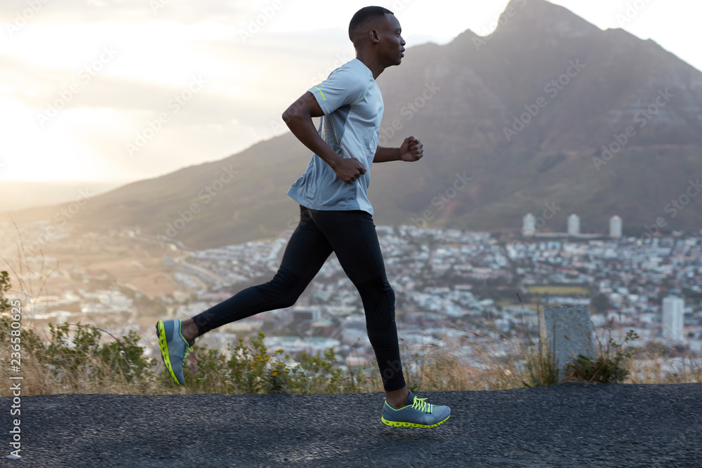 Sideways shot of handsome active man runs against mountain beautiful scenery, photographed in motion, enjoys workout, being very fast and energetic, wears sportclothes. Athlete black male outside