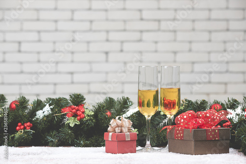 Champagne glasses set next to a pretty red gift box on table with pine branches and christmas ornaments, white brick wall blurred background for copy space holizon lay out photo