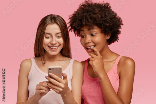 Indoor shot of curious cheerful rwo multiethnic women read news in internet via cell phone, make purchases online, stands closely, have interracial relationships, isolated over pink background. photo