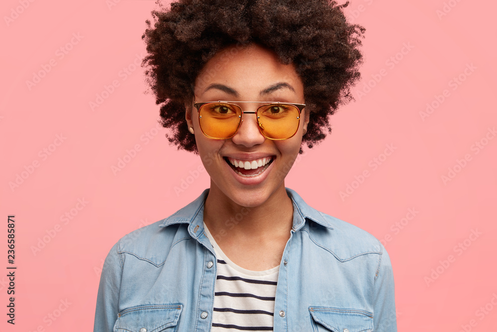 Close up shot of female hipster with black skin, curly hair, smiles positively, wears shades and denim shirt, rejoices positive moment in life, happy to make shopping during weekend, isolated on pink