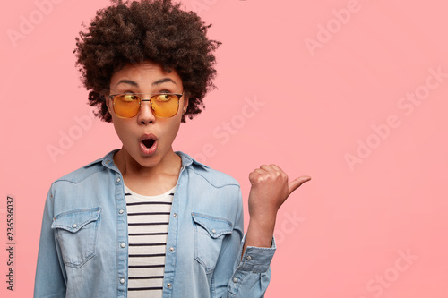 Stunned emotive young woman with unexpected reaction points aside as notices famous person at street, being surprised, wears trendy shades and denim jacket, models against pink studio background