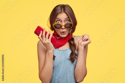 Photo of surprised emotive dark haired woman in trendy shades holds cell phone, hears something astonishing, wears red bandana, models over yellow background. People, reaction and style concept.