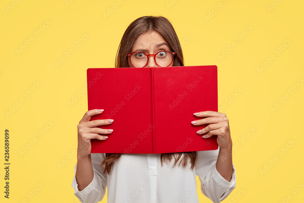 Puzzled dark haired woman raises eyebrows, looks with puzzlement over red textbook, wears white shirt, models against yellow wall, learns new information, prepares home assignment, reads story Puzzled dark haired woman raises eyebrows, looks with puzzlement over red textbook, wears white shirt, models against yellow wall, learns new information, prepares home assignment, reads story