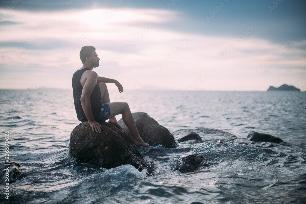 A young man looks at the sunset by the sea. A handsome guy is sitting on the rocks in the sea at sunset.