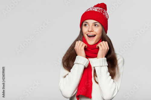 Surprised happy  woman looking sideways in excitement. Christmas girl wearing knitted warm hat and scarf  isolated on gray background
