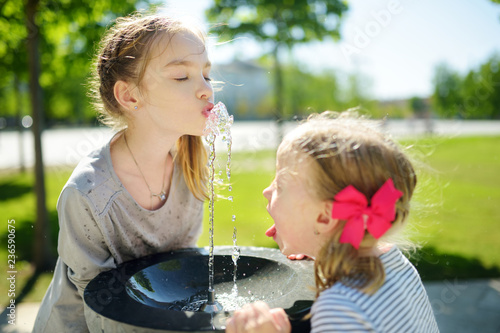Obraz na plátně Two sisters having fun with drinking water fountain on warm and sunny summer day
