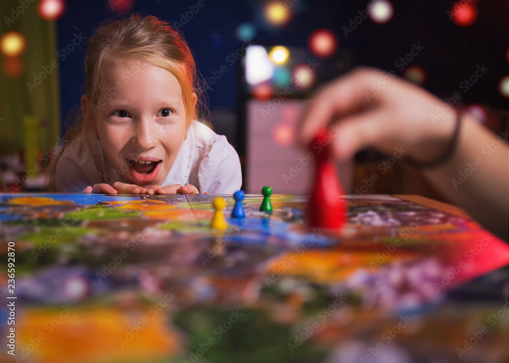 Board game concept - your move. Little girl watched the game and shock from the action move. Board game field, many figures. Kid girl play in board game at home on dark blurred background