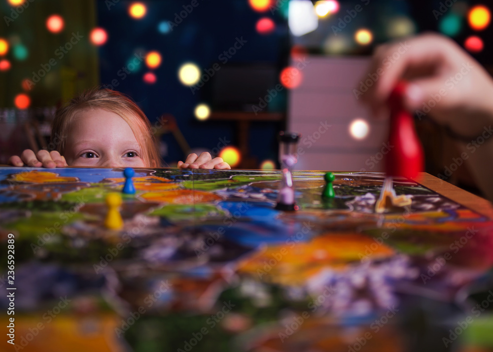 Board game concept - your move. Little girl watched the game and shock from the action move. Board game field, many figures. Kid girl play in board game at home on dark blurred background Board game concept - your move. Little girl watched the game and shock from the action move. Board game field, many figures. Kid girl play in board game at home on dark blurred background