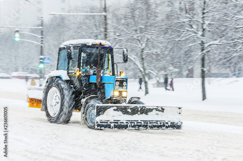 Blue snow plow tractor removes snow from the city road. Snowstorm in the city - seasonal abstract motion blur background