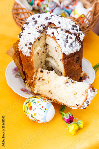 Easter cake with white and black chocolate sauce, spring flowers and an egg on a bright yellow tablecloth, behind an Easter basket