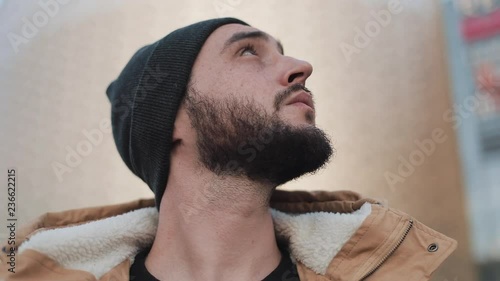 Young beard man tourist walk on city travelling in Europe standinh near shopping mall looks up at the city. He is wearing an autumn jacket and knitted hat. Close up photo