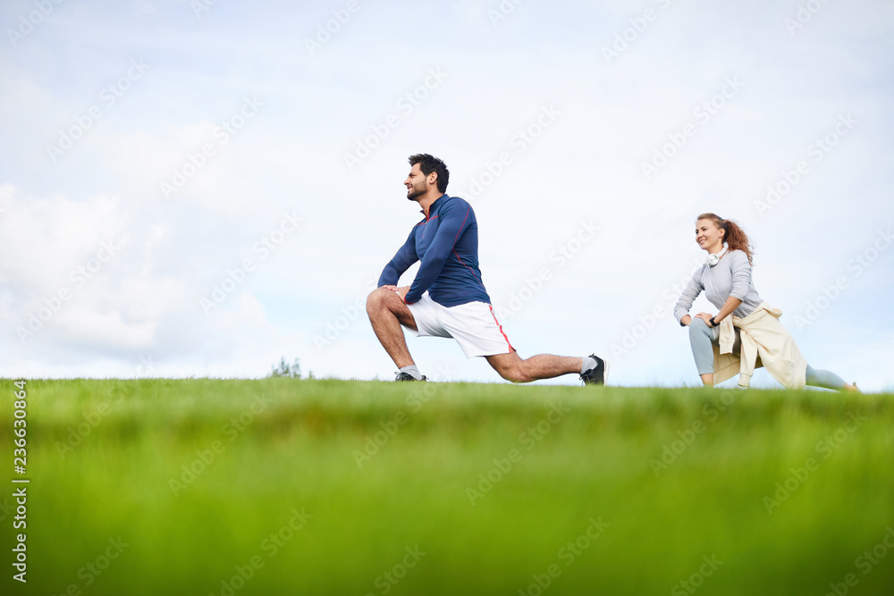 Young active man and woman in sportswear bending knees and stretching legs during outdoor workout Young active man and woman in sportswear bending knees and stretching legs during outdoor workout