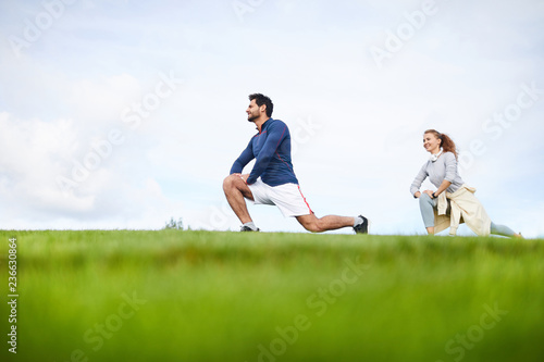 Young active man and woman in sportswear bending knees and stretching legs during outdoor workout © pressmaster