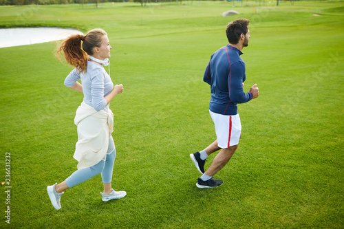 Active young man and woman running down green grass in rural environment or in park