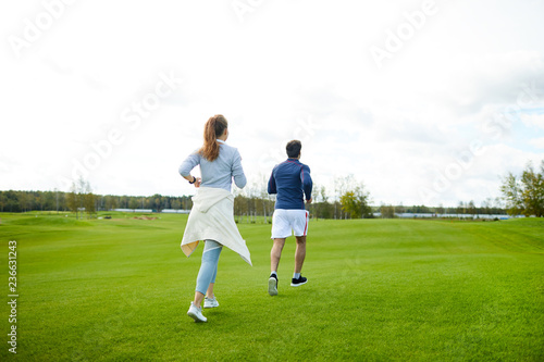 Rear view of active girl and guy in sportswear running down green grass in the morning