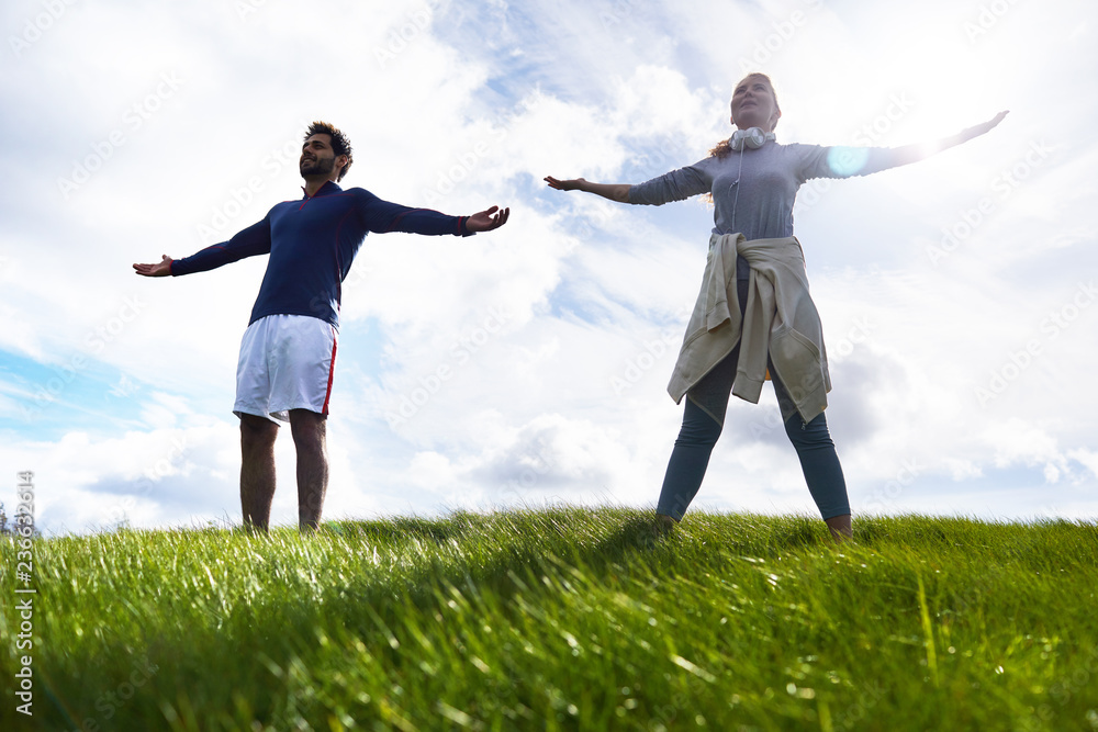 Young couple in activewear standing on green field with their arms outstretched Young couple in activewear standing on green field with their arms outstretched