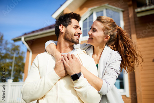 Happy young woman embracing her husband on background of their new house