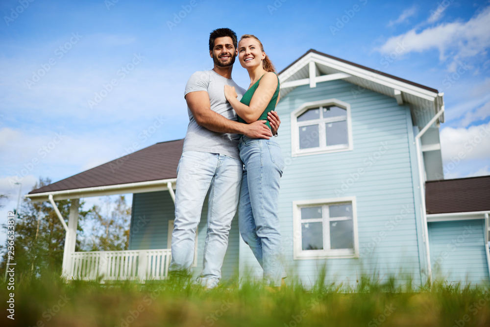 Young man and woman standing in embrace on green lawn by their new house Young man and woman standing in embrace on green lawn by their new house