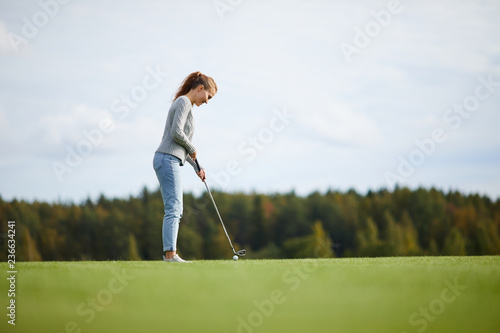 Young active woman in casualwear standing on golf field and getting ready to hit ball with club