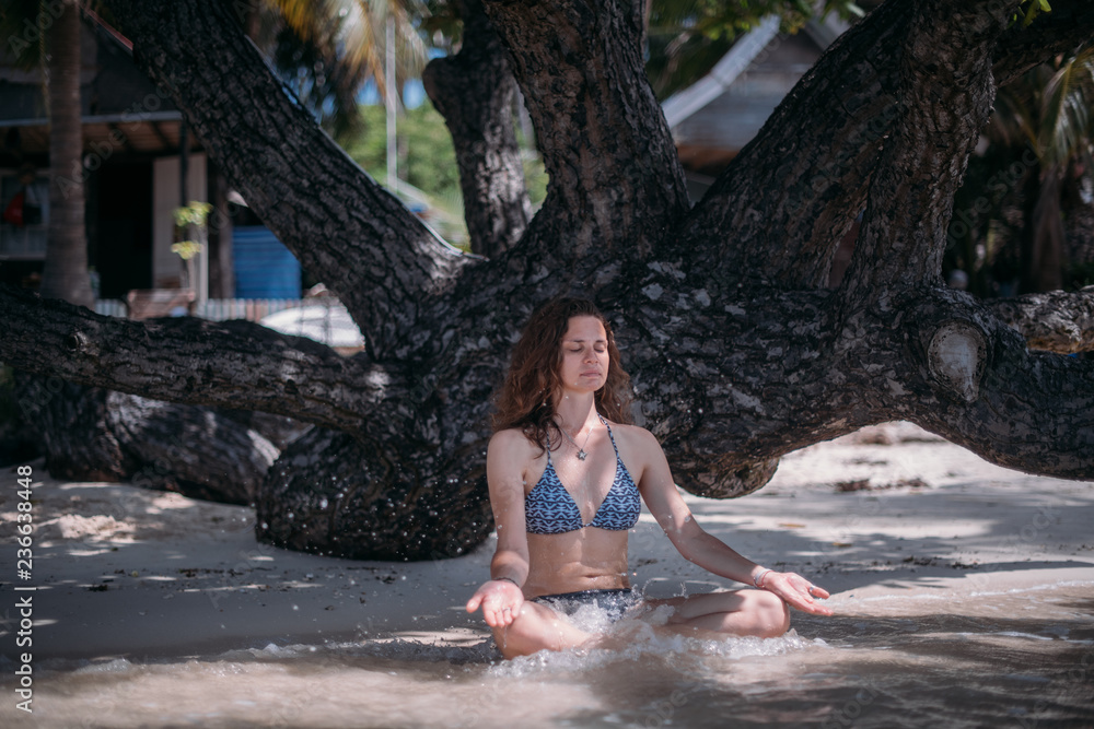 Fototapeta premium Young slim girl sitting in the shade of a tree in the sea waves on the coast of a tropical island