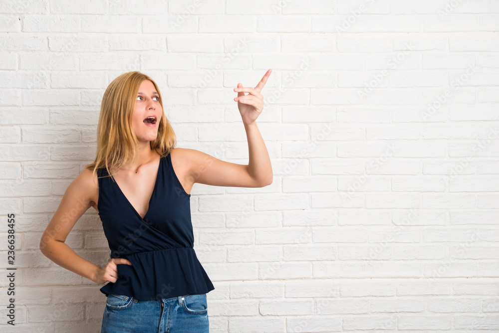Blonde woman pointing finger to the side and presenting a product while smiling in a confident pose on white brick wall background Blonde woman pointing finger to the side and presenting a product while smiling in a confident pose on white brick wall background